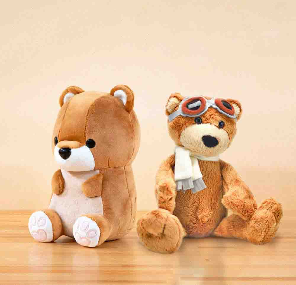 Two teddy bear plush toys on a wooden surface, one with a simple design and the other wearing aviator goggles and a scarf.