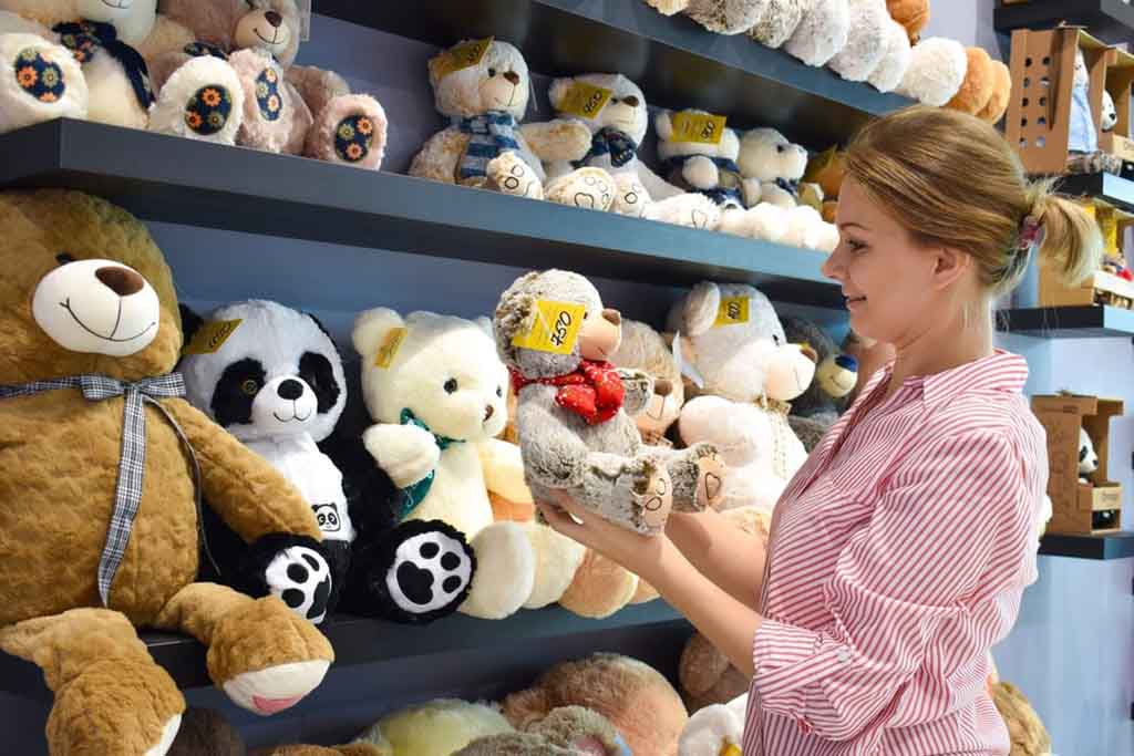 Smiling woman in a striped shirt holding a teddy bear while browsing a store shelf full of plush toys, including pandas and other bears.