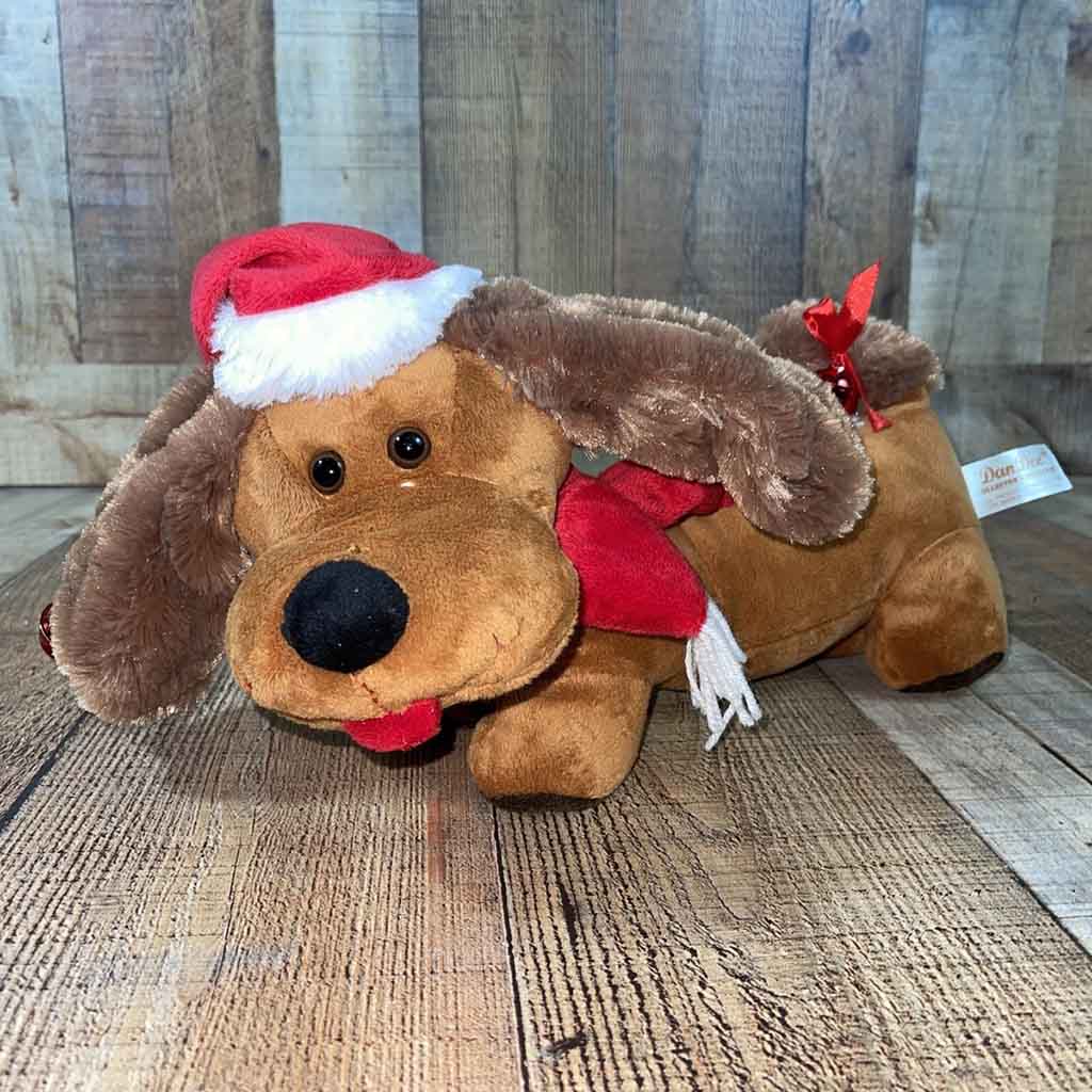 Brown plush dog wearing a red Santa hat and scarf, lying on a wooden floor with floppy ears and a festive look.