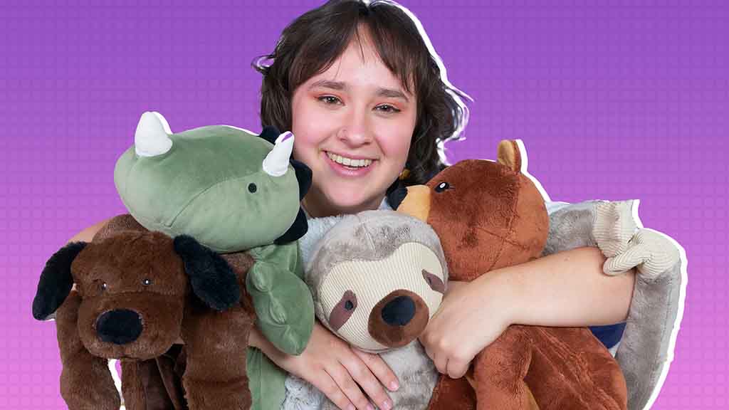 Young girl smiling while hugging multiple stuffed animals including a dog, dinosaur, sloth, and bear against a purple background.
