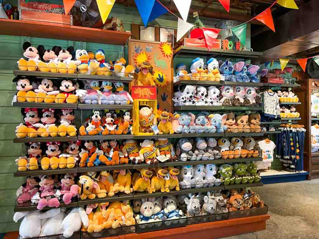 Colorful shelves filled with Disney plush toys including Mickey, Minnie, Donald, Goofy, Stitch, Dumbo, and Simba inside a Disney store.
