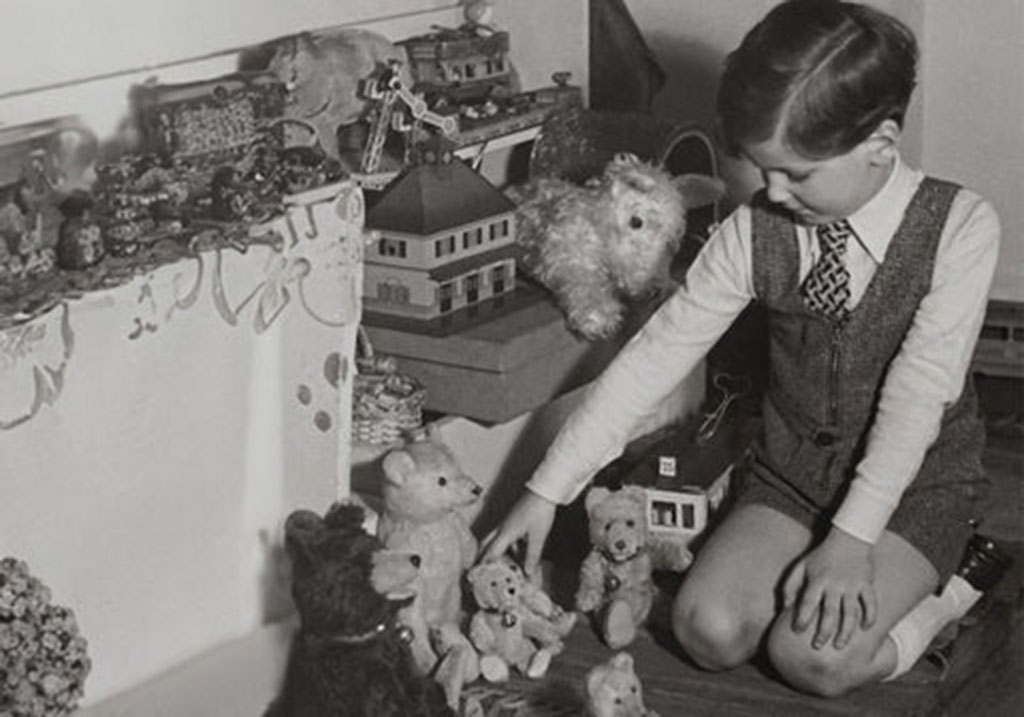 Black and white photo of a young boy in formal attire playing with teddy bears and toy animals beside a decorated table and dollhouse.