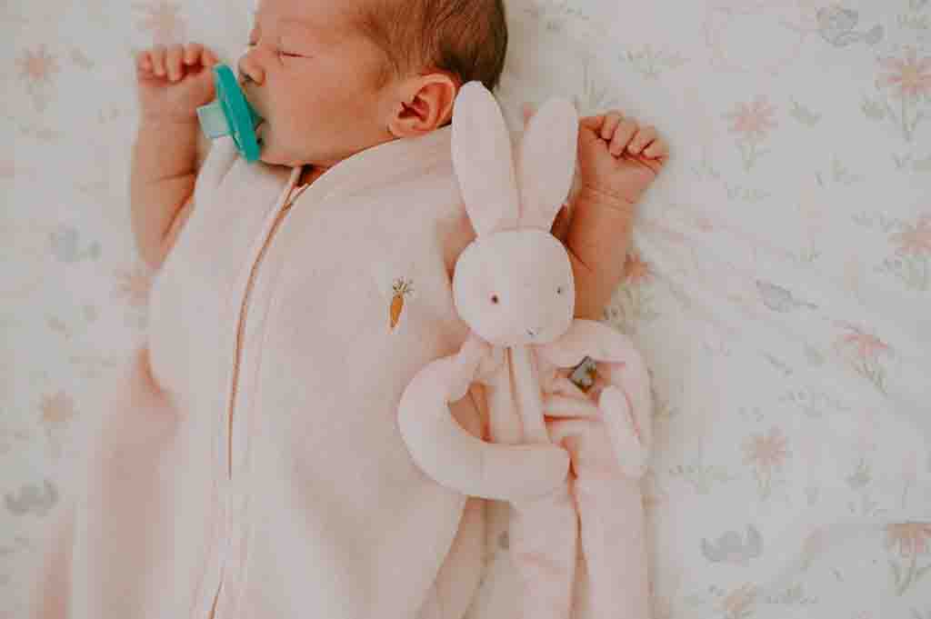 Infant sleeping peacefully with a pacifier, dressed in a pink sleep sack, holding a soft bunny comfort plush toy.