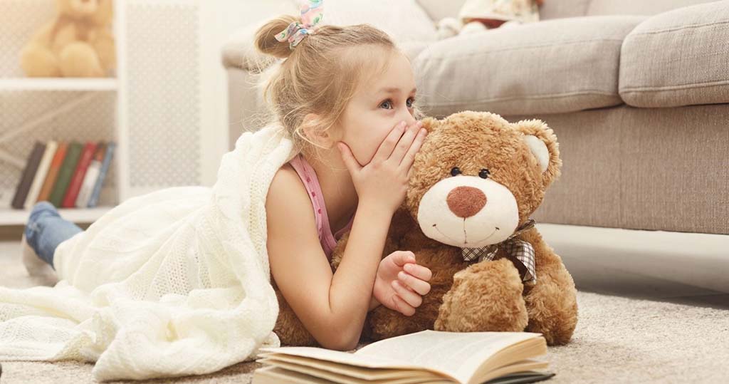 Little girl lying on the carpet with a blanket, whispering to her teddy bear while reading a book at home.