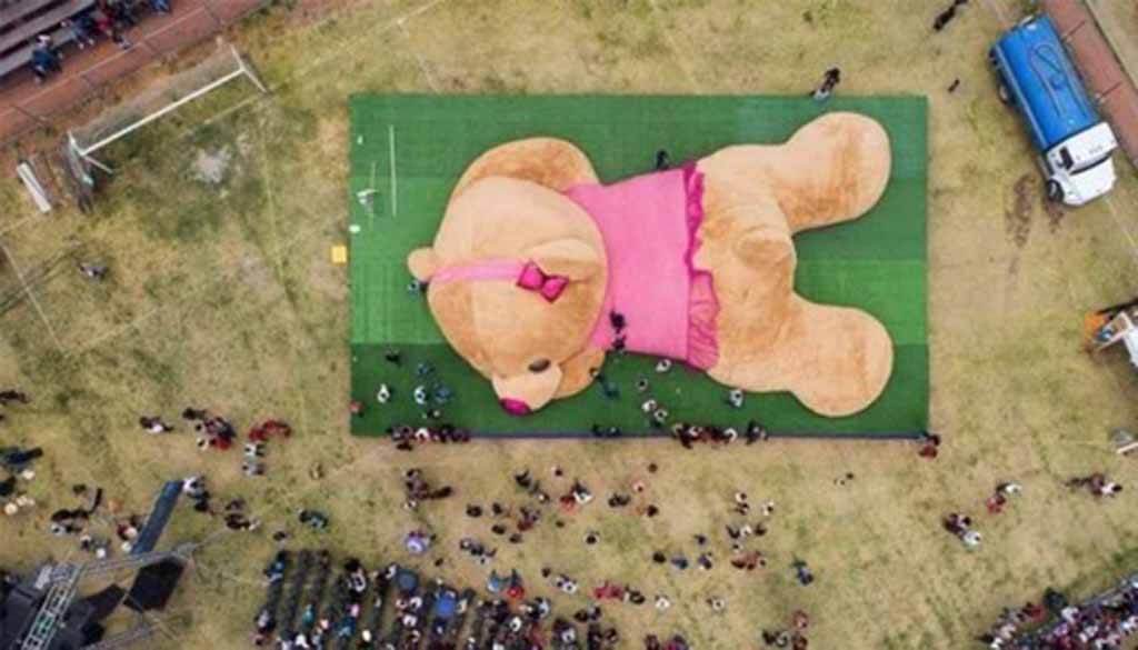 Giant teddy bear in pink outfit displayed outdoors with people gathered around.