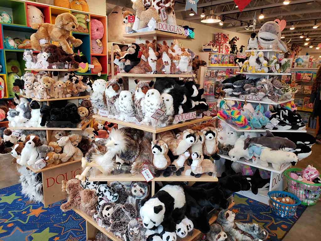 Shelves filled with a wide variety of plush toys including dogs, owls, pandas, unicorns, and sharks in a colorful toy store.