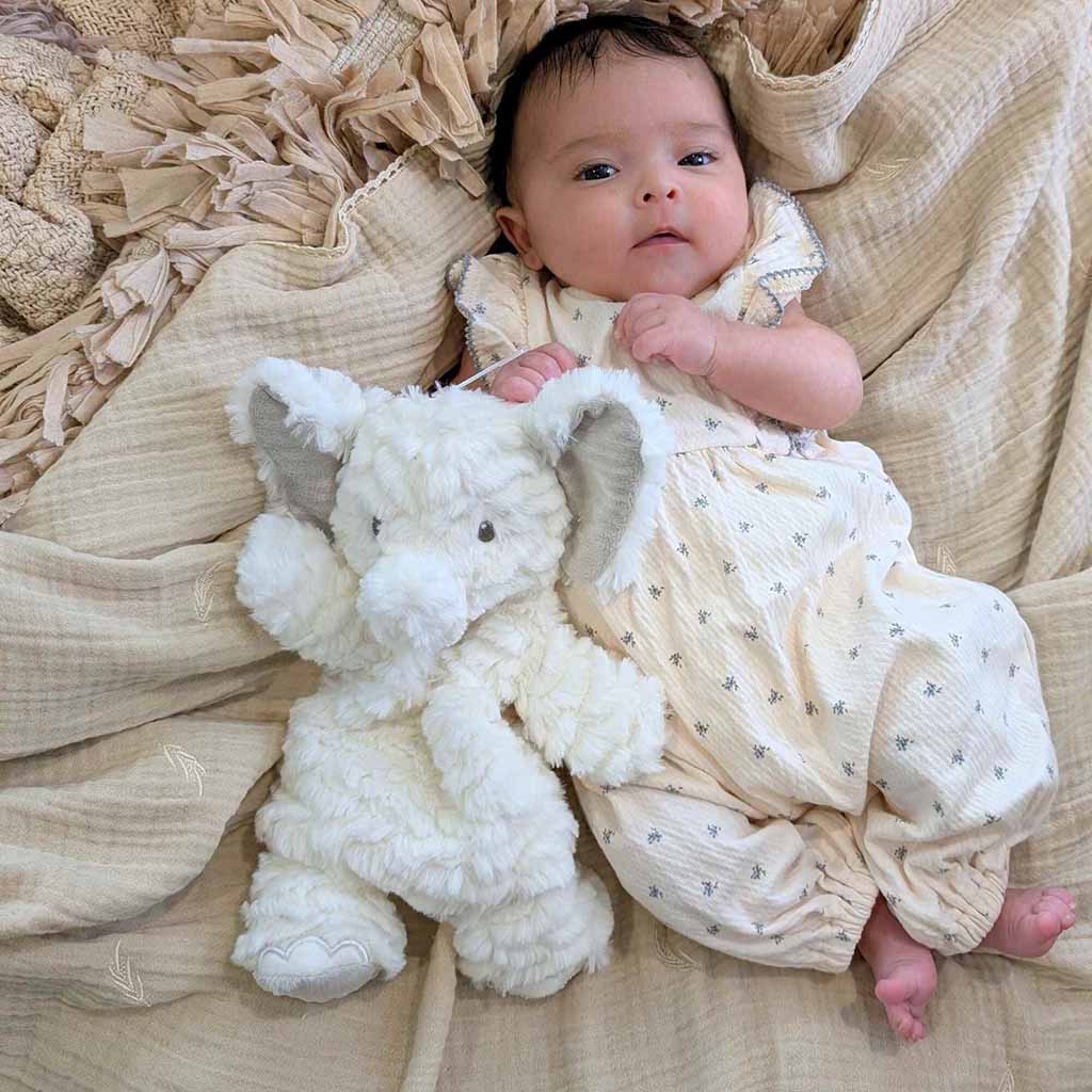Infant lying on soft blankets while holding a fluffy white elephant stuffed toy.