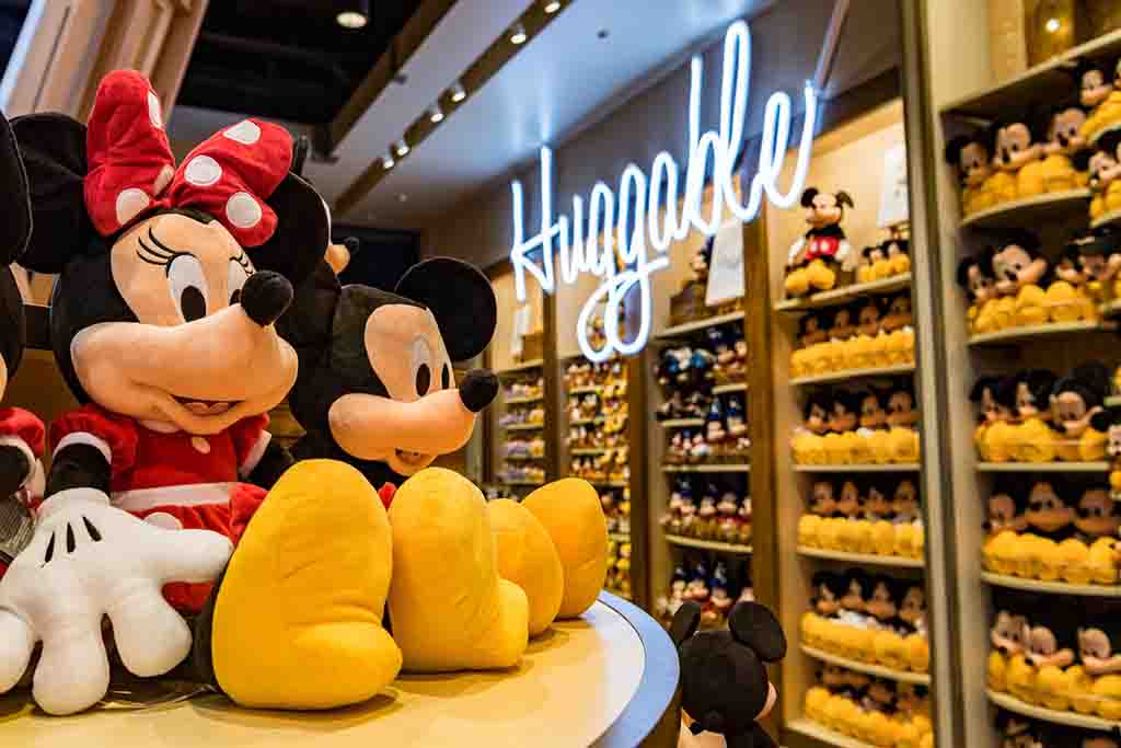 Large Mickey and Minnie plush toys displayed in a Disney store with shelves full of plushies and a glowing “Huggable” sign above.