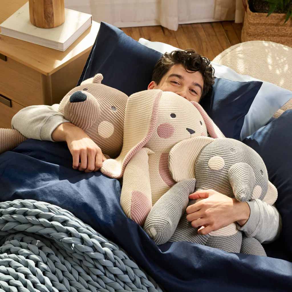 A man lying in bed hugging three large knitted plush toys—a bear, a rabbit, and an elephant—while wrapped in a navy blue blanket.