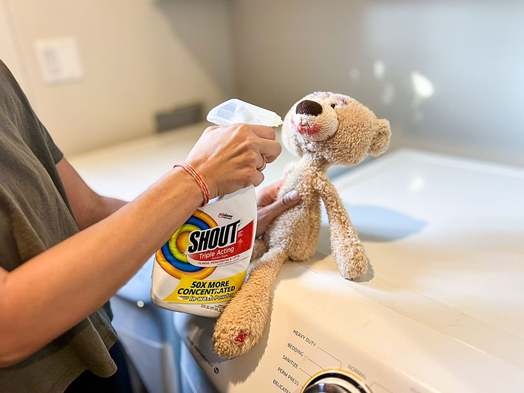 Person spraying stain remover on a dirty teddy bear toy placed on top of a washing machine.