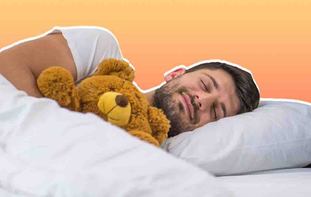 Adult man peacefully sleeping in bed while hugging a brown teddy bear against a white pillow and blanket.