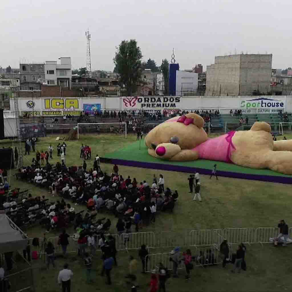 Giant teddy bear in pink dress displayed in stadium with large crowd of people gathered around.