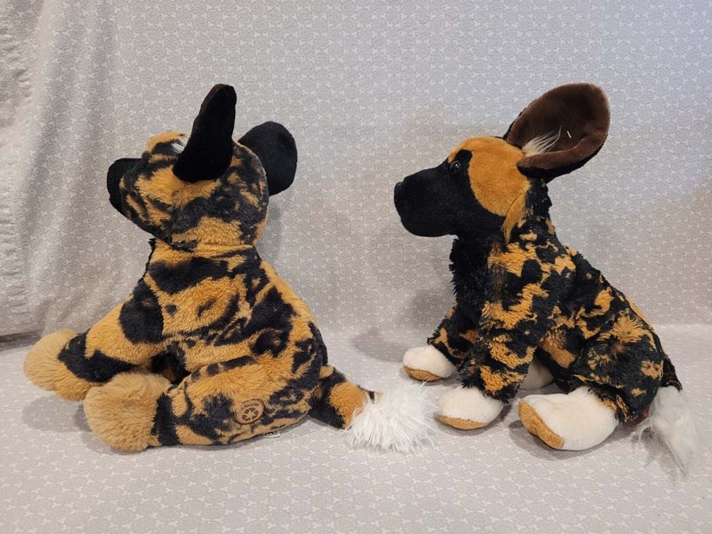 Two African wild dog plush toys with realistic black, brown, and tan fur patterns, sitting side by side on a light patterned background.