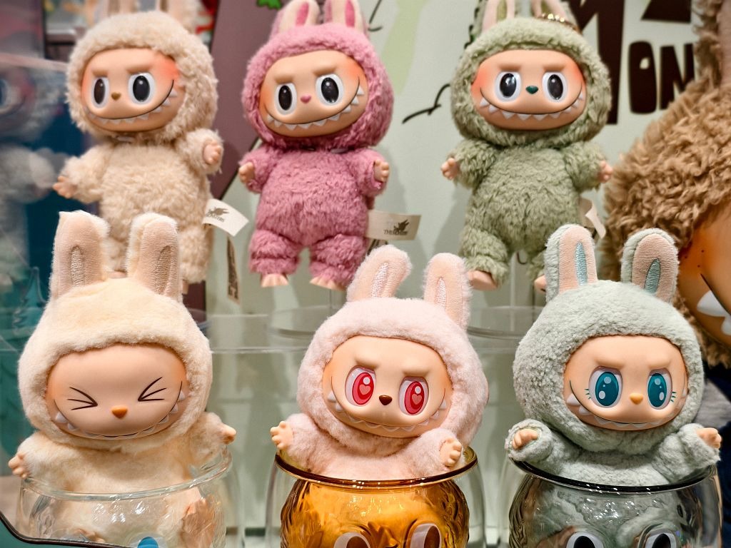 Colorful plush dolls dressed in fluffy animal costumes with bunny ears, displayed in a shop with expressive faces and playful designs.
