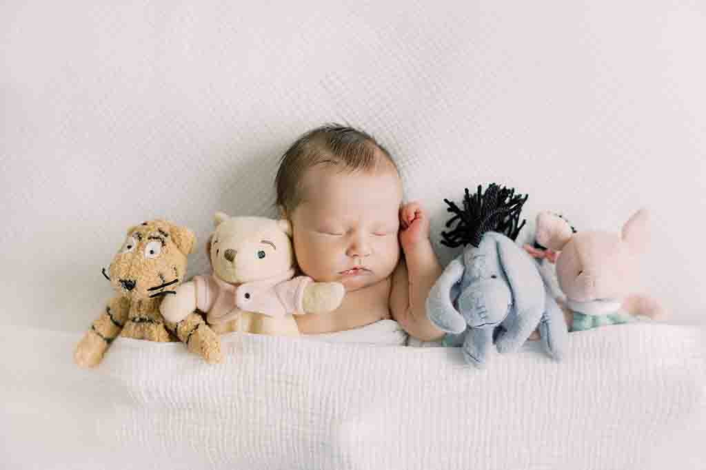 Baby peacefully asleep under a white blanket, surrounded by plush toys including a tiger, bear, donkey, and piglet.