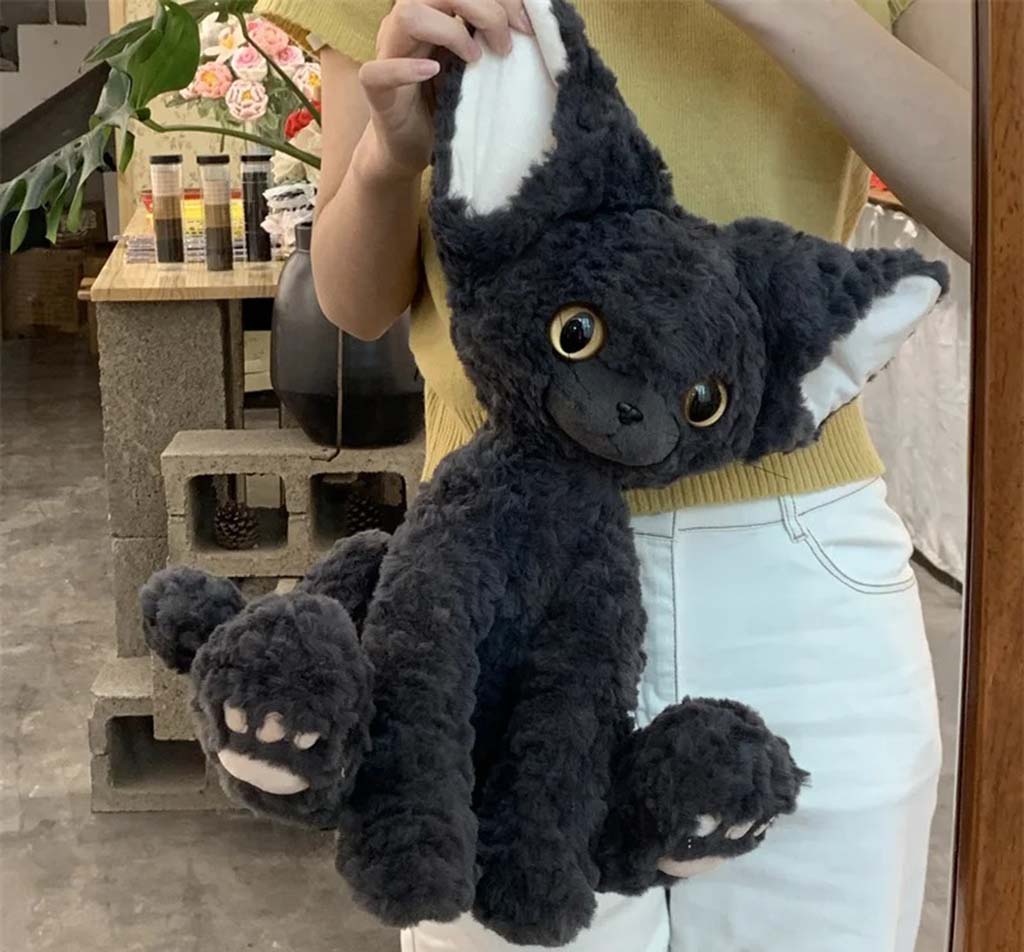 Person holding a fluffy black cat plush toy with oversized ears, golden eyes, and soft paw details, indoors with plants and shelves in the background.