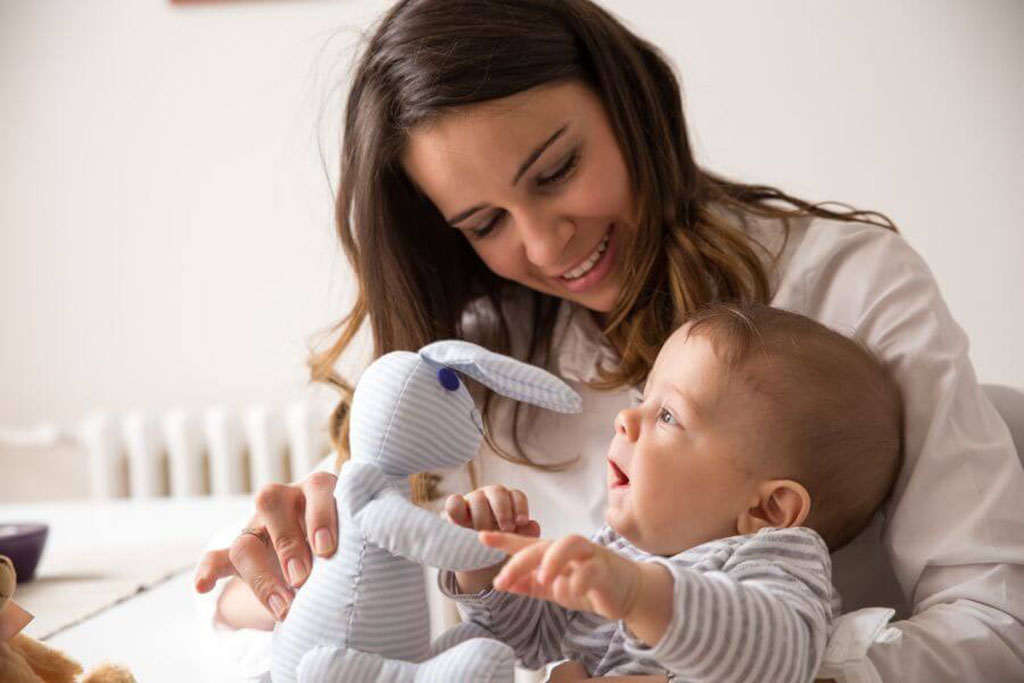 Smiling mother holding her baby as they play with a striped soft bunny plush toy, creating a joyful bonding moment.