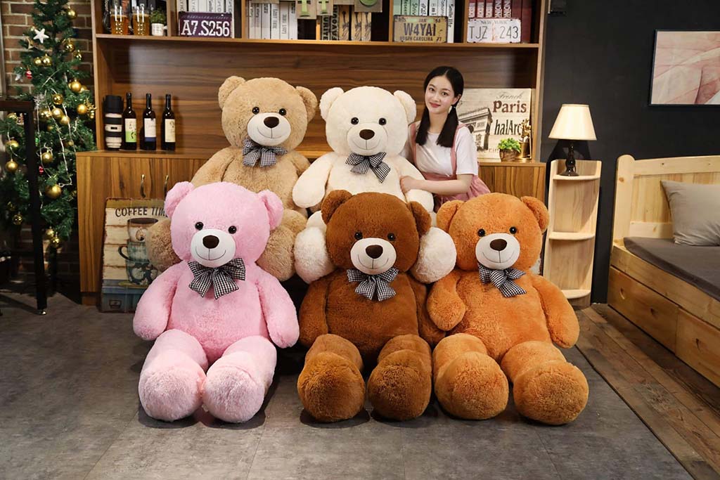 Young woman sitting with a group of oversized teddy bears in different colors, including pink, brown, white, and beige, displayed in a cozy room setting.