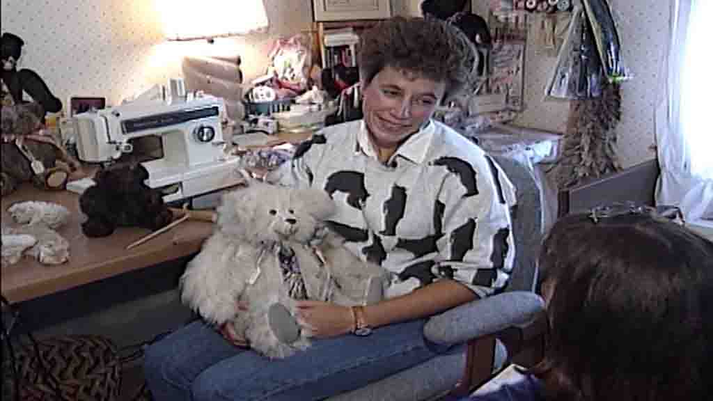 Woman smiling while holding a handmade teddy bear in a sewing room, with a sewing machine and plush materials on the table.