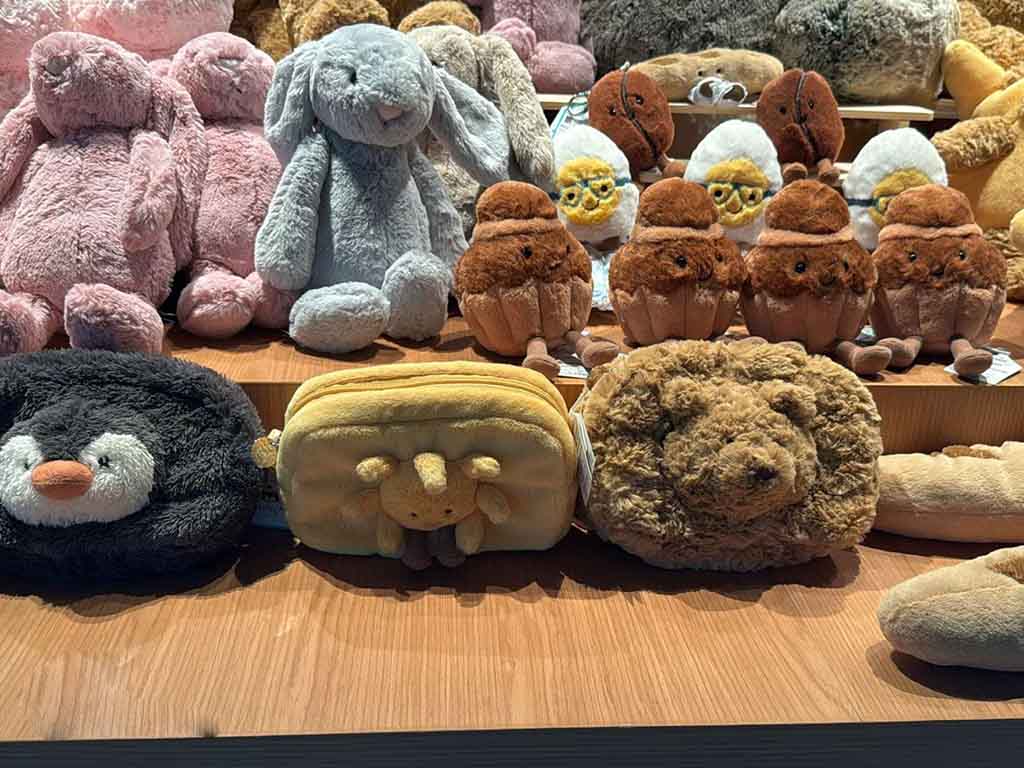A collection of soft plush toys displayed on wooden shelves, including penguins, bunnies, bears, and other animal designs in pastel and earthy tones.