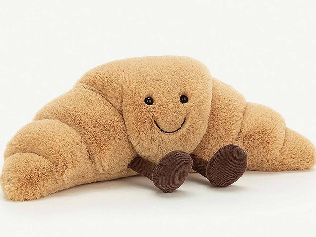 Cute croissant-shaped plush toy with a smiling face and brown corduroy feet on a plain white background.