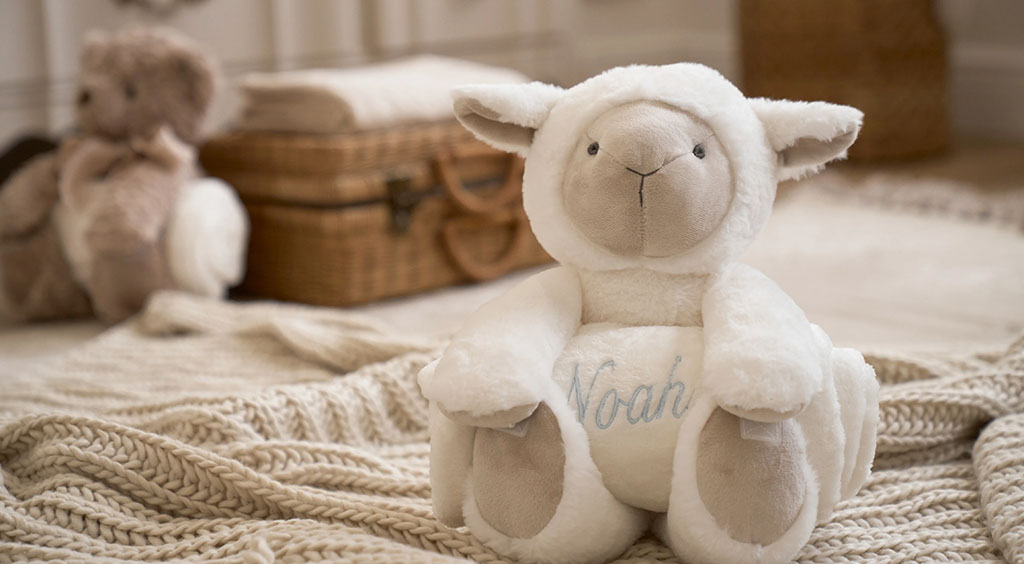 Soft white plush lamb with “Noah” embroidered on its belly, sitting on a knitted blanket in a warm nursery setting.