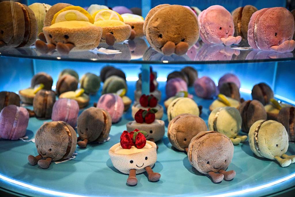Cute dessert-themed plush toys shaped like macarons and cakes with smiling faces, displayed on illuminated blue shelves.