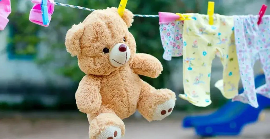 A brown teddy bear hanging on a clothesline with colorful clothespins beside baby clothes drying outdoors.