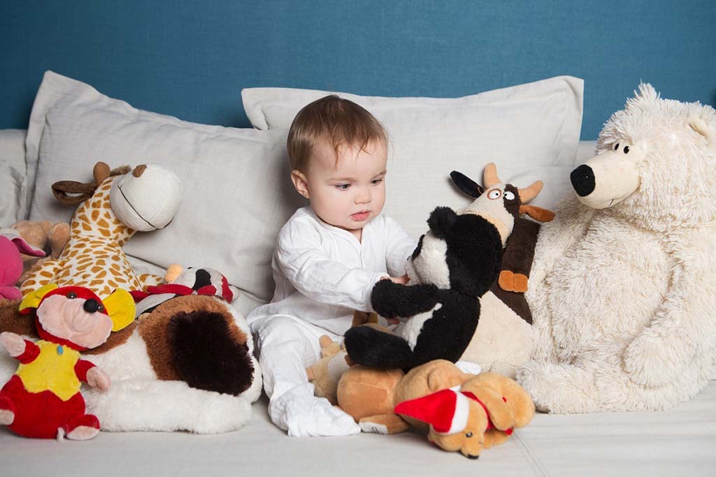 Toddler in white pajamas sitting on a bed surrounded by colorful plush toys including a giraffe, bear, and cow.