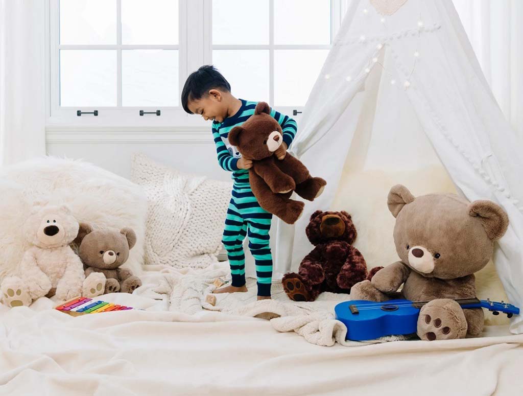 A young child in striped pajamas playing with teddy bears inside a cozy bedroom tent surrounded by plush toys, a blue guitar, and soft blankets.