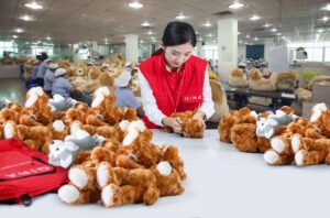 A quality inspector in a red vest examines teddy bears in a plush toy factory, with workers assembling toys in the background.