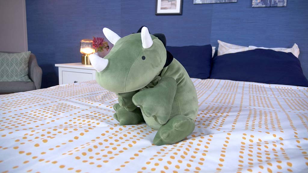 Soft green rhinoceros stuffed animal with white horns sitting on a neatly made bed with patterned sheets and navy pillows in a cozy bedroom.