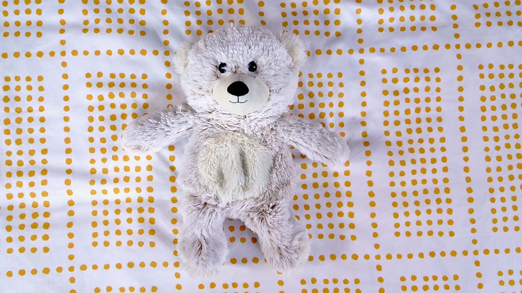 Light gray teddy bear with cream belly lying on a white and yellow dotted bedsheet, showing a cozy and cute plush toy design.