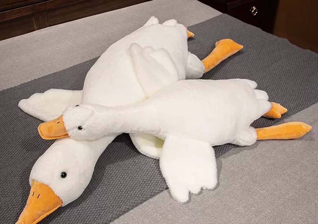 Two large white goose plush toys with orange beaks and feet lying on a gray blanket in a cozy bedroom setting.