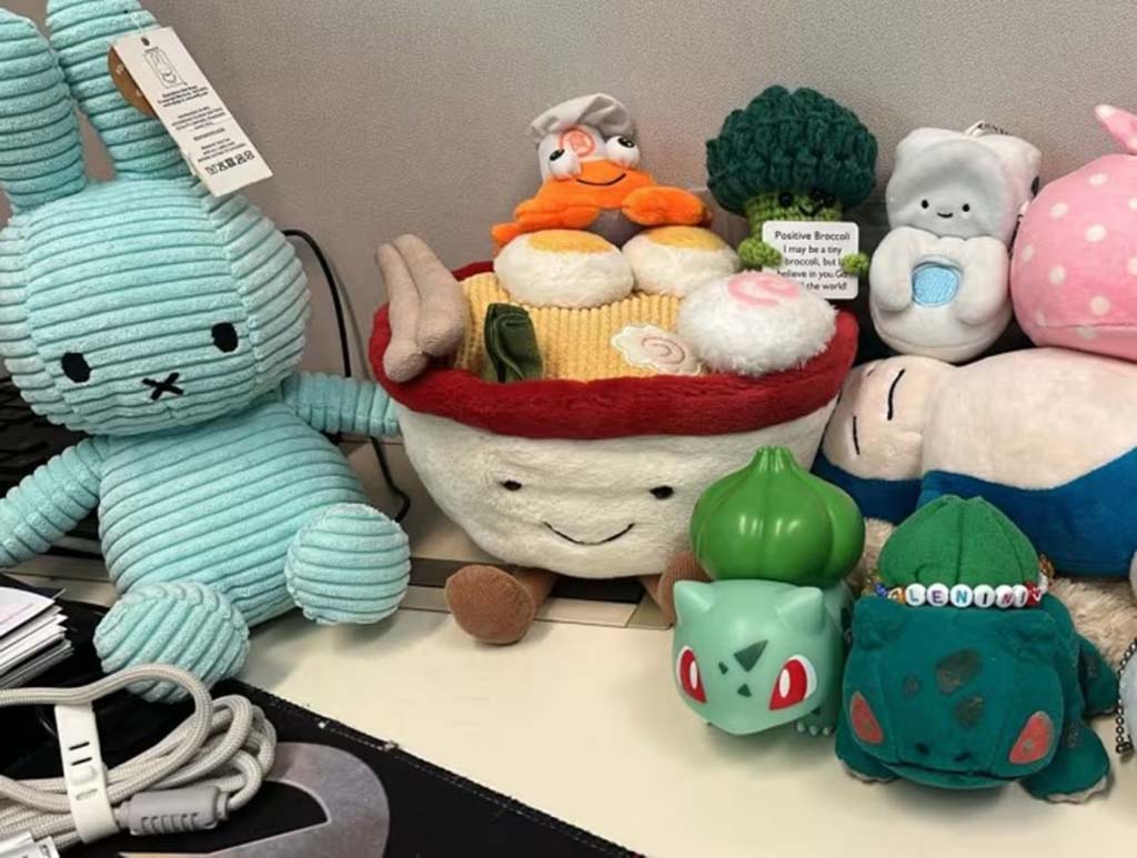 A group of colorful plush toys arranged on a desk, including a blue bunny, a ramen bowl plush, broccoli, and characters from Pokémon.