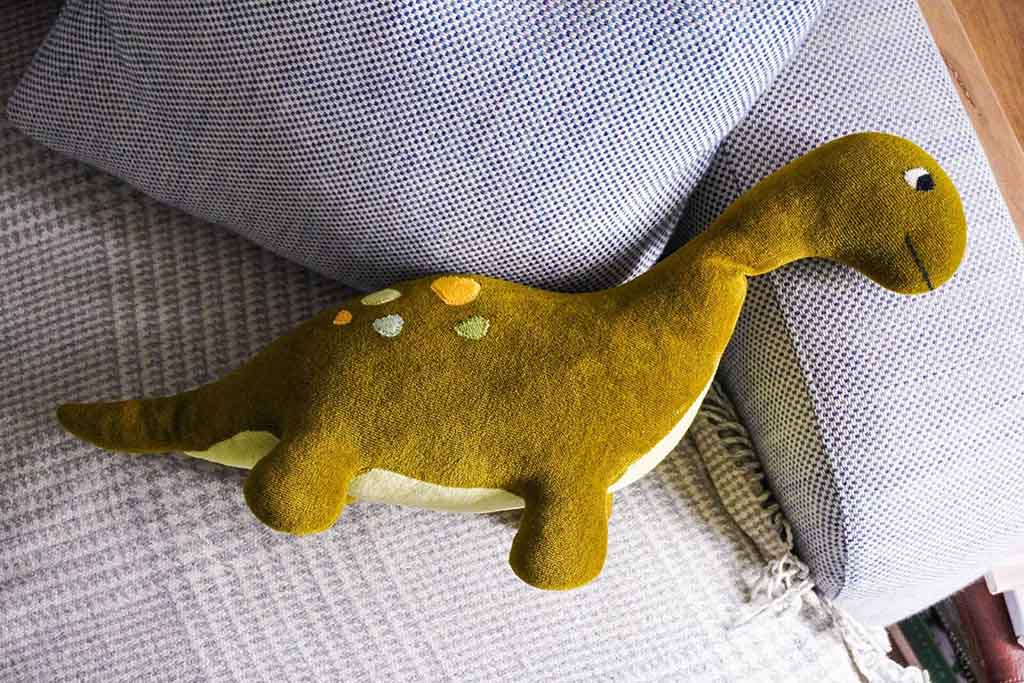 Soft green dinosaur plush toy with embroidered details lying on a gray fabric sofa cushion.