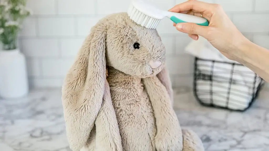 Hand brushing a beige plush bunny toy with a soft brush on a marble surface, showing gentle cleaning and maintenance for stuffed animals.