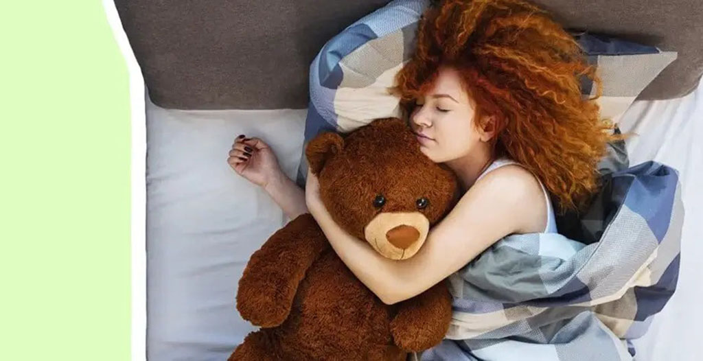A young woman with curly red hair peacefully sleeping in bed while hugging a large brown teddy bear under a blue plaid blanket.