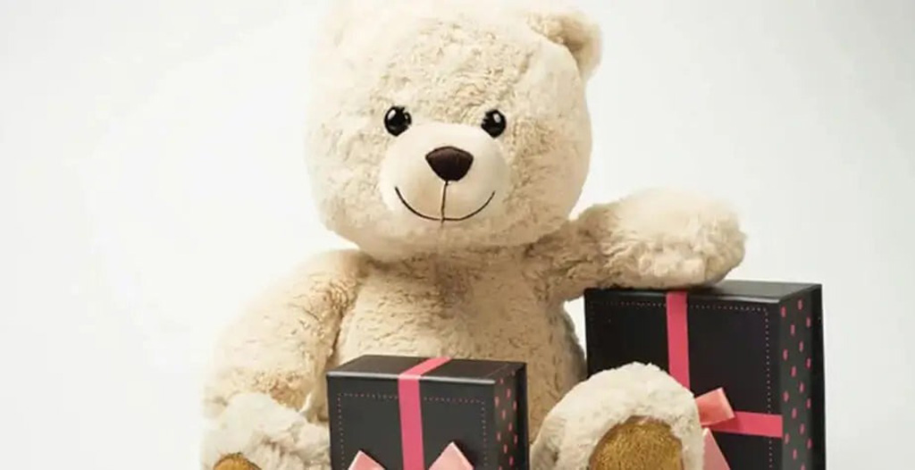 Cream-colored teddy bear sitting beside two black gift boxes tied with pink ribbons on a light background.