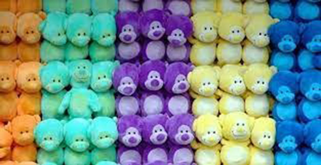 Rows of plush teddy bears in gradient rainbow colors including orange, green, purple, yellow, and blue, neatly stacked together.