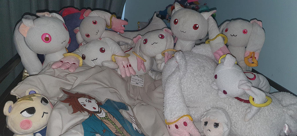 A bed covered with multiple white and pink anime-style plush toys featuring large red eyes and ring-shaped accessories, along with other character plushies and blankets.