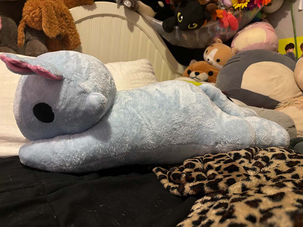 Light blue plush toy with pink ears lying on a bed, surrounded by other stuffed animals and soft pillows in a cozy bedroom setting.