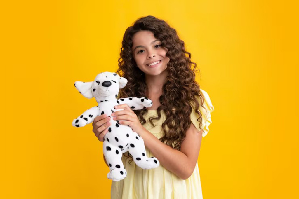 A cheerful young girl with long curly hair wearing a yellow dress, smiling and holding a white Dalmatian plush toy against a bright yellow background.