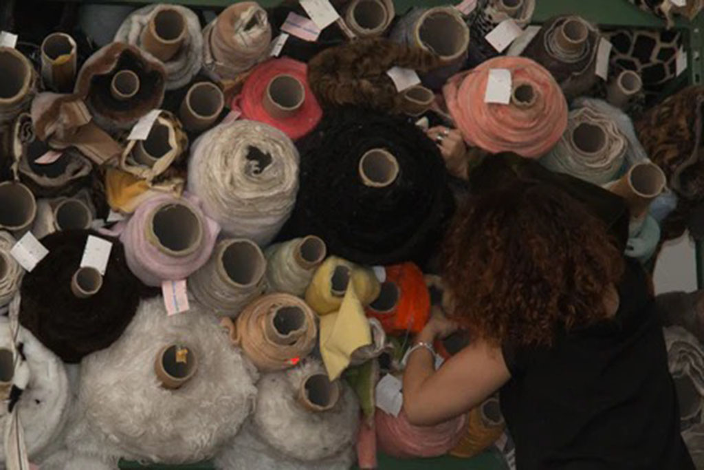 Person selecting from a variety of plush fabric rolls in different colors and textures stored on shelves in a toy manufacturing workshop.