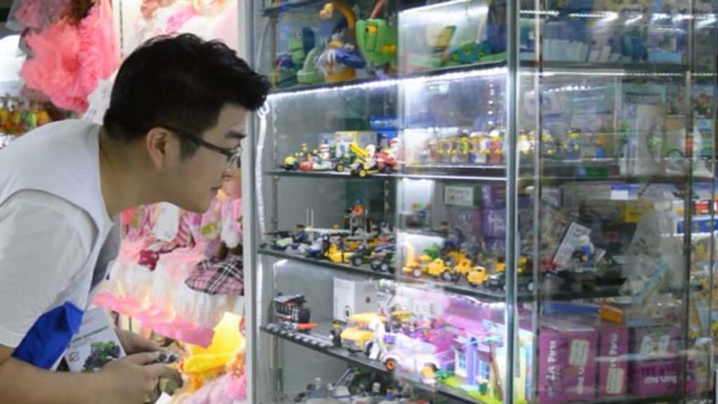 Man inspecting toy display shelves filled with miniature cars, action figures, and playsets in a brightly lit toy market.