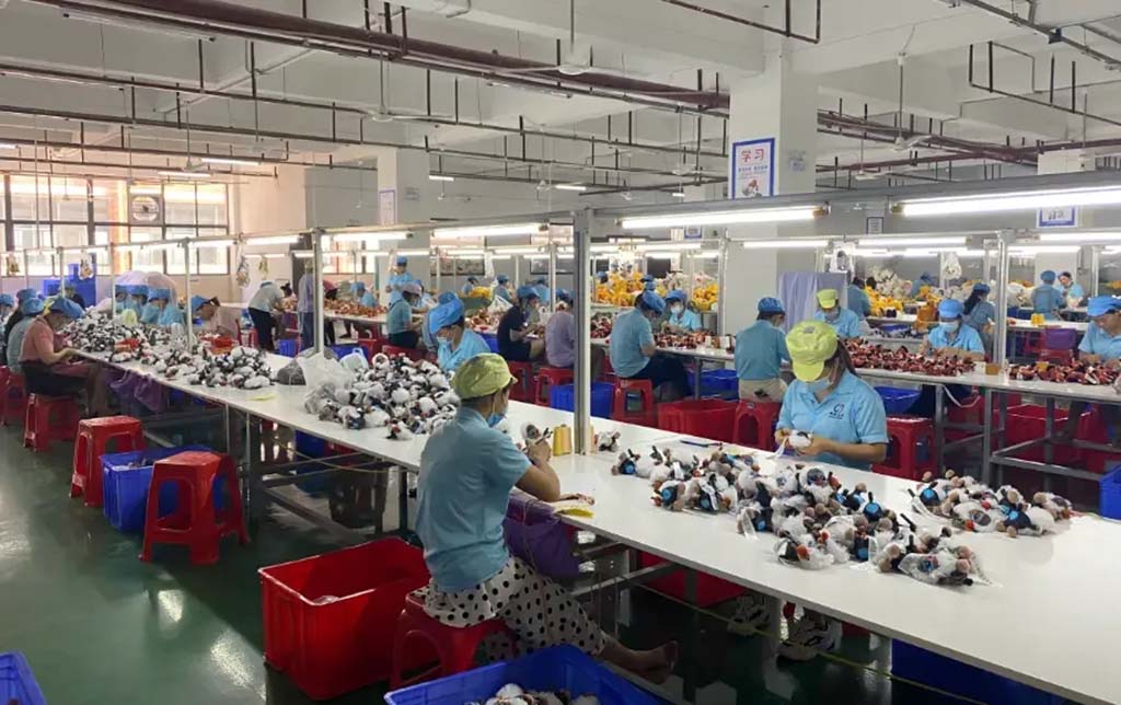 Factory workers in blue uniforms assembling and inspecting plush toys at long production tables under bright lighting in a clean manufacturing facility.