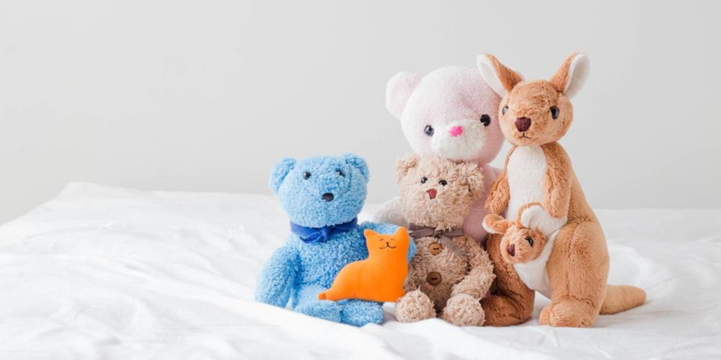 A group of soft plush toys including a blue bear, brown bear, white bear, kangaroo with joey, and small orange cat plush sitting together on a white bed background.