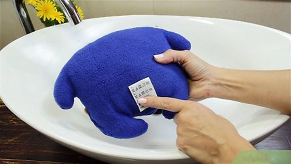Person holding a blue plush toy over a white sink while reading its fabric care label before cleaning.
