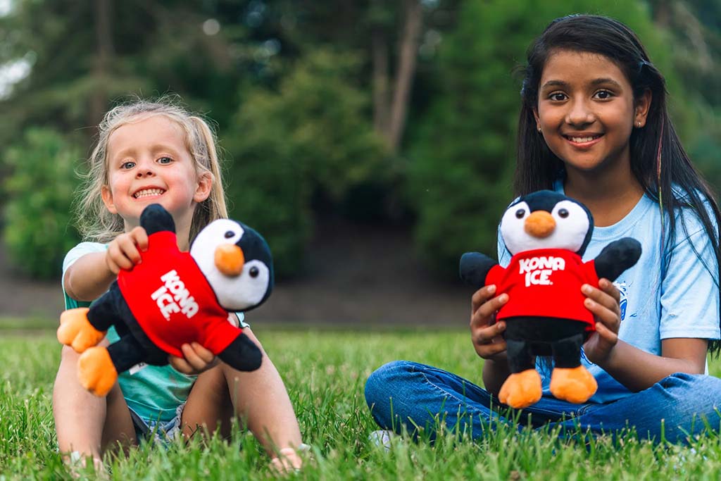 Two smiling children sitting on grass in a park, each holding a black and white penguin plush toy wearing a red "Kona Ice" shirt, enjoying a sunny day outdoors.