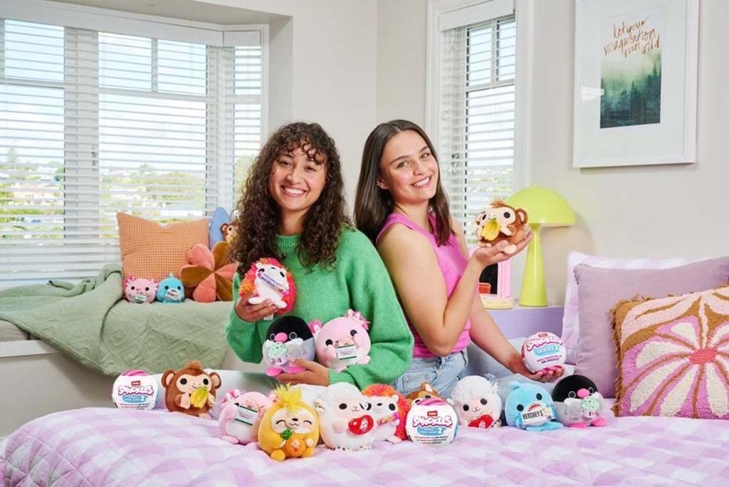Two smiling girls sitting on a bed surrounded by colorful round plush toys, holding their favorites in a bright, cozy bedroom filled with sunlight and decor.